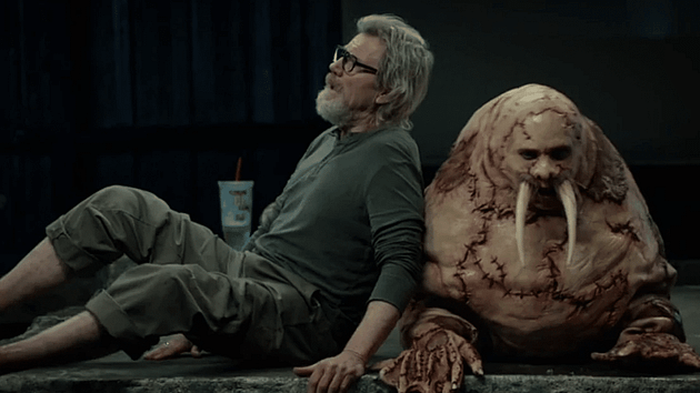 A middle-aged man with glasses and gray hair sitting next to a monster-like creature with tusks, both appearing relaxed in a dimly lit environment.