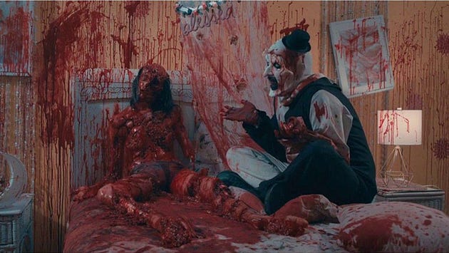 A clown sitting on a bed covered in blood, interacting with a lifeless, bloody figure propped up beside him in a room with splattered walls.