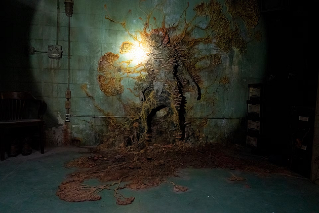 A shadowy figure partially blended into a mossy wall in a dimly lit room, with organic growths surrounding it.
